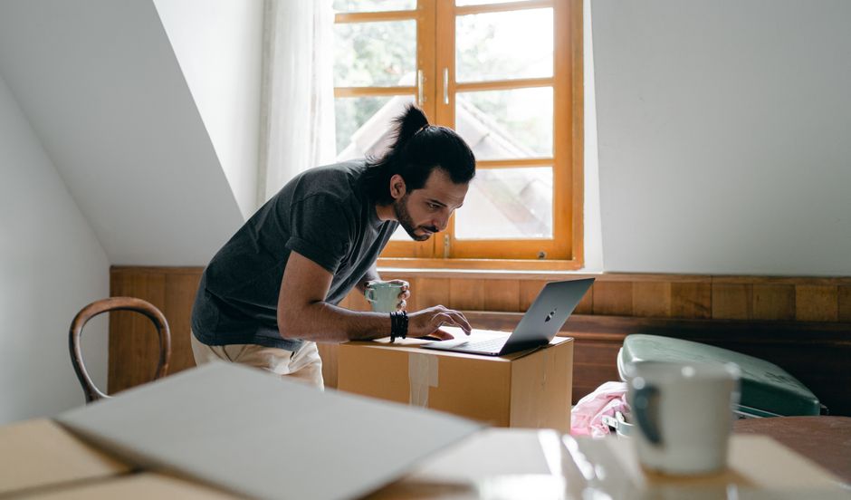 Side view of focused bearded male in casual outfit with cup of hot drink browsing laptop while packing stuff into carton boxes