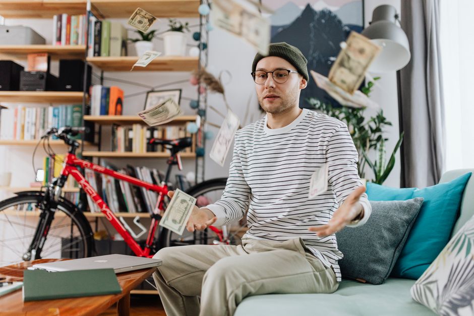 Young man tossing money in a cozy room, showcasing indoor leisure and wealth.